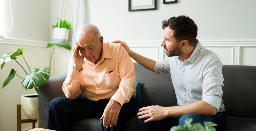 Älterer Mann shat einen Schlaganfall und sitzt mit starken Kopfschmerzen auf dem Sofa. Daneben sitzt sein Sohn und versucht mit dem Vater zu sprechen.